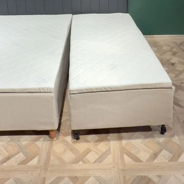 Två färdigmonterade sängmoduler står bredvid varandra på ett ljust trägolv med en smal springa emellan. Varje modul har ett eget beige överdrag, en egen quiltad madrass och egna ben.