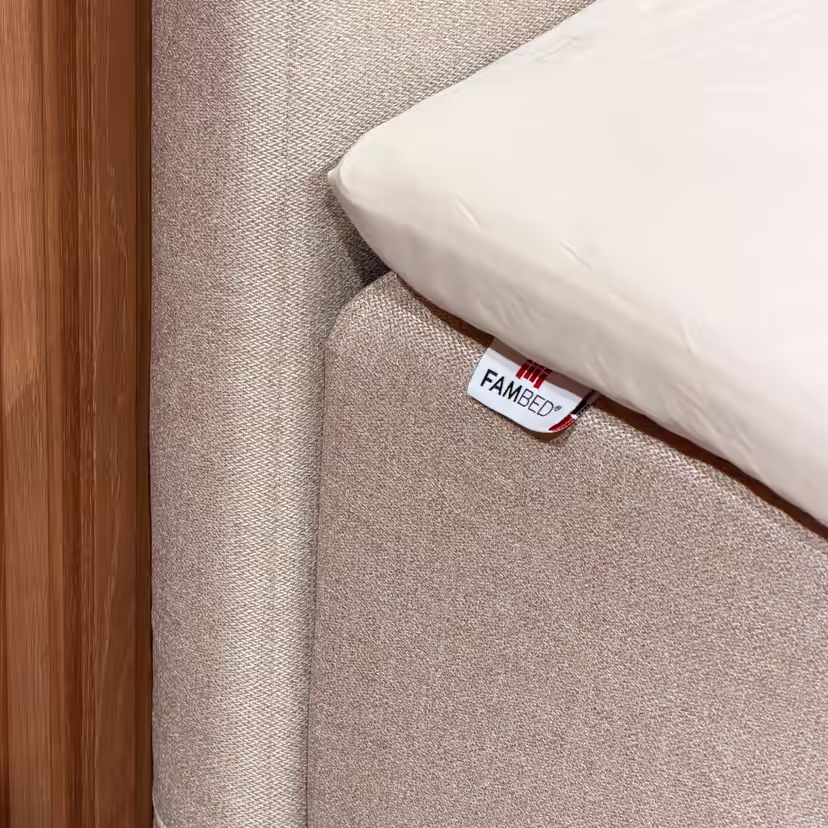 Närbild av det beige överdraget runt sängens hörn, med FAMBED-märke och ett vitt lakan ovanpå